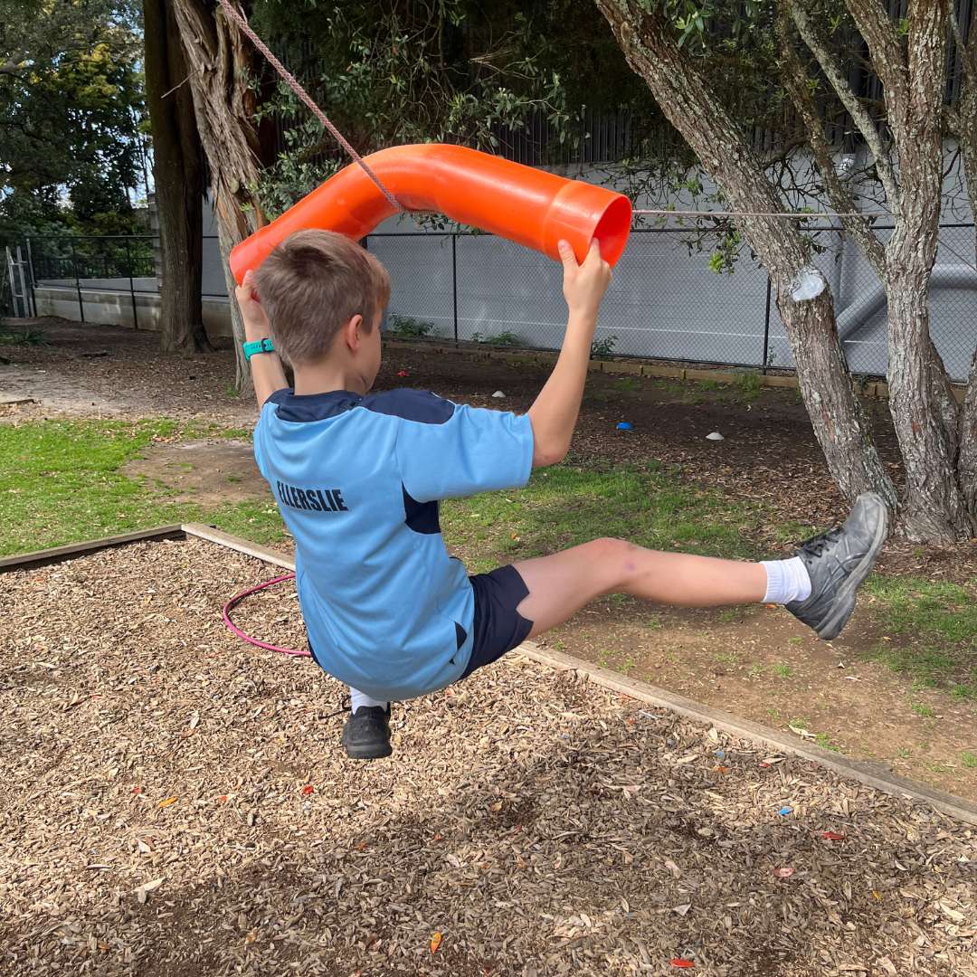 Child in blue shirt swinging on an orange plastic tube attached to a tree in a playground.