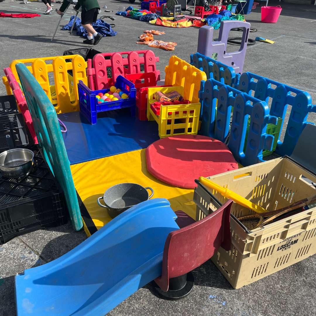 Enclosed outdoor play area with colourful plastic fencing, blue slide, chairs, balls, and toys.