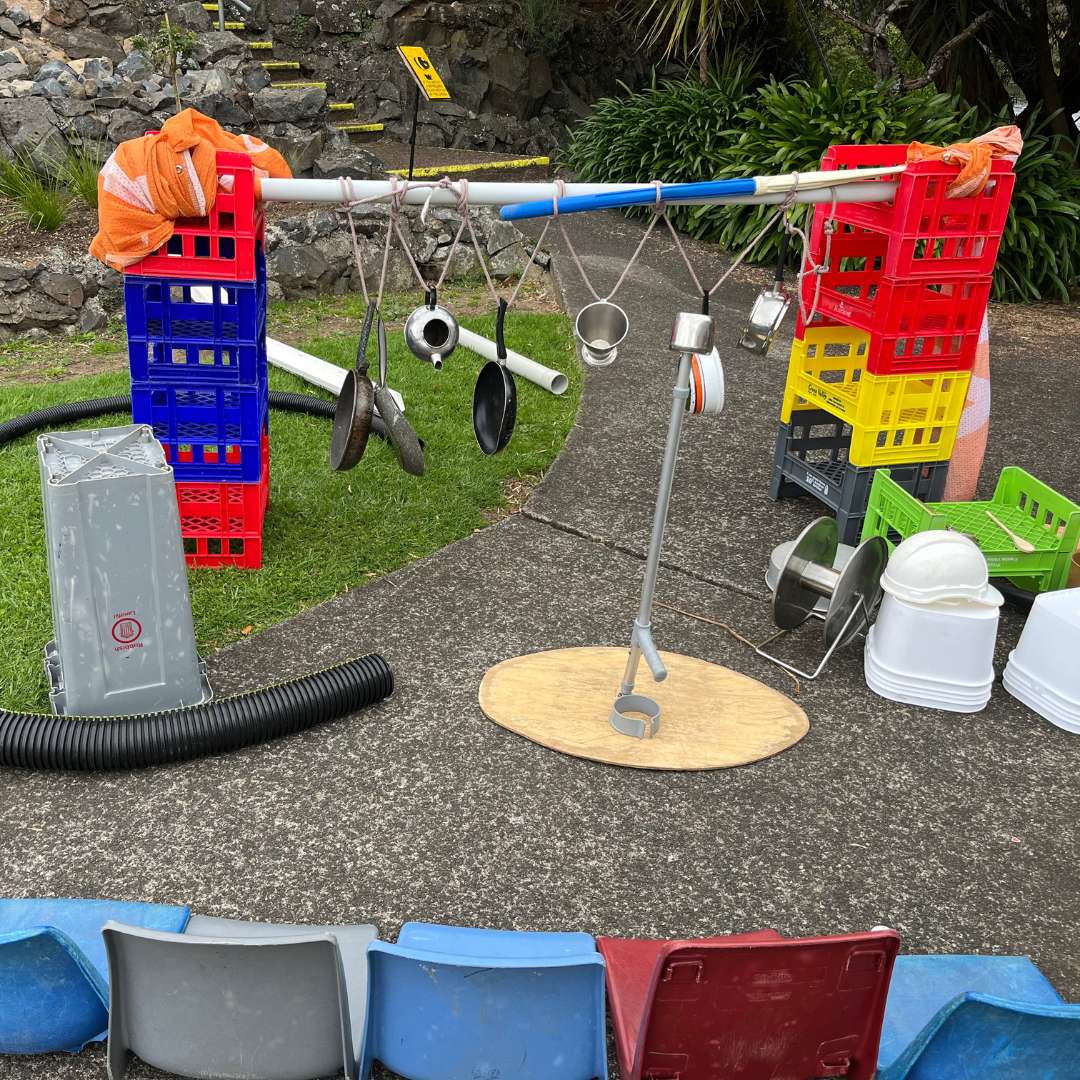 Outdoor kitchen utensils hanging from a pole supported by plastic crates with chairs and various containers nearby.